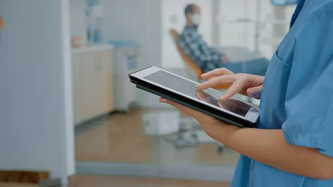 Prontuário odontológico. Dentista consultando prontuário odontológico em tablet com paciente na cadeira ao fundo.